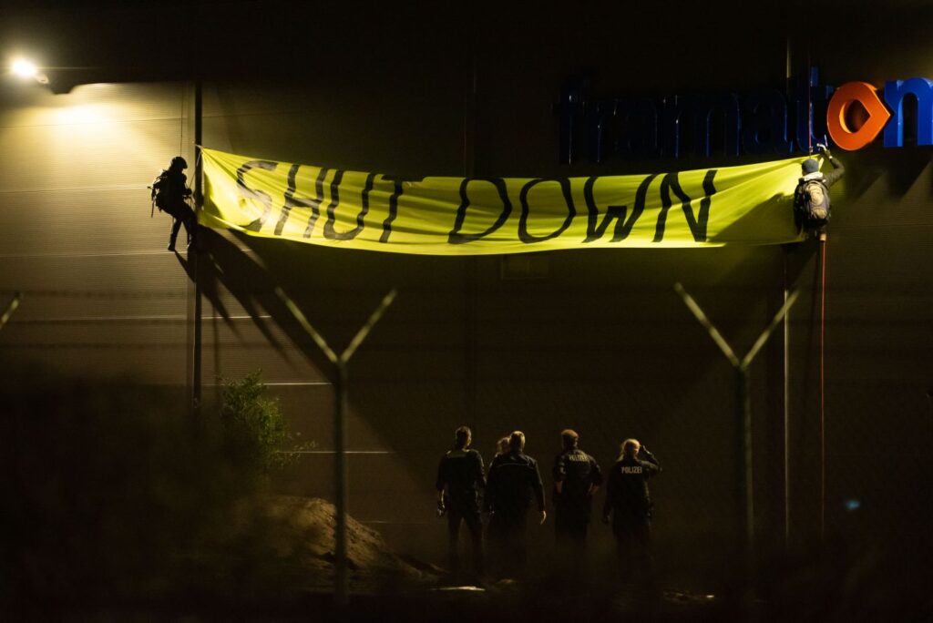 Großes Banner mit Aufschrift "SHUT DOWN" im Dunkeln an Gebäudefassade, Menschen hängen daneben an Seilen, unten steht Polizei