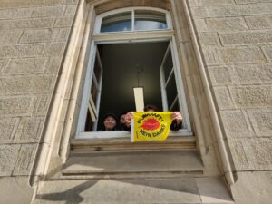 Aus einem geöffneten hohen Fenster eines Steingebäudes schauen mehrere Menschen und halten eine Atomkraft-Nein-Danke-Fahne