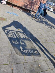 Auf dem Boden ist der Schatten einer Person zu sehen, welche ein Transparent hält, bei der die Worte "Shut down - auch die Repression" ausgeschnitten sind.