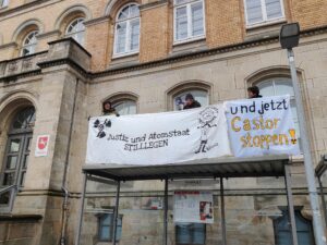 3 Menschen stehen auf einer Bushaltestelle vorm Landgericht Osnabrück und halten zwei Transparente mit den Aufschriften "Justiz und Atomstaat stilllegen" und "Und jetzt Castor stoppen"