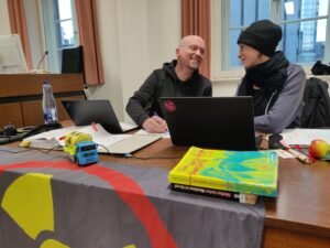 Zwei Menschen sitzen an einem Tisch, vor ihnen Bücher und ein kleiner nachgebauter Casotr und lachen sich an.